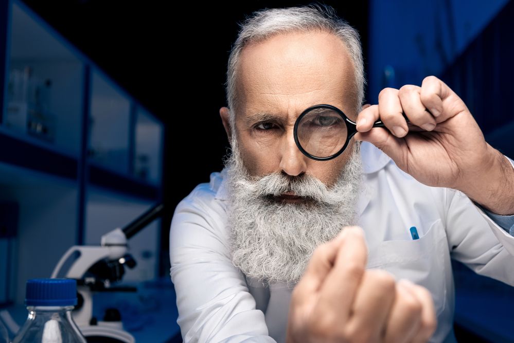 Cient&iacute;fico con lupa inspecciona minuciosamente en un laboratorio, rodeado de equipo especializado; enfoque en investigaci&oacute;n y precisi&oacute;n cient&iacute;fica.