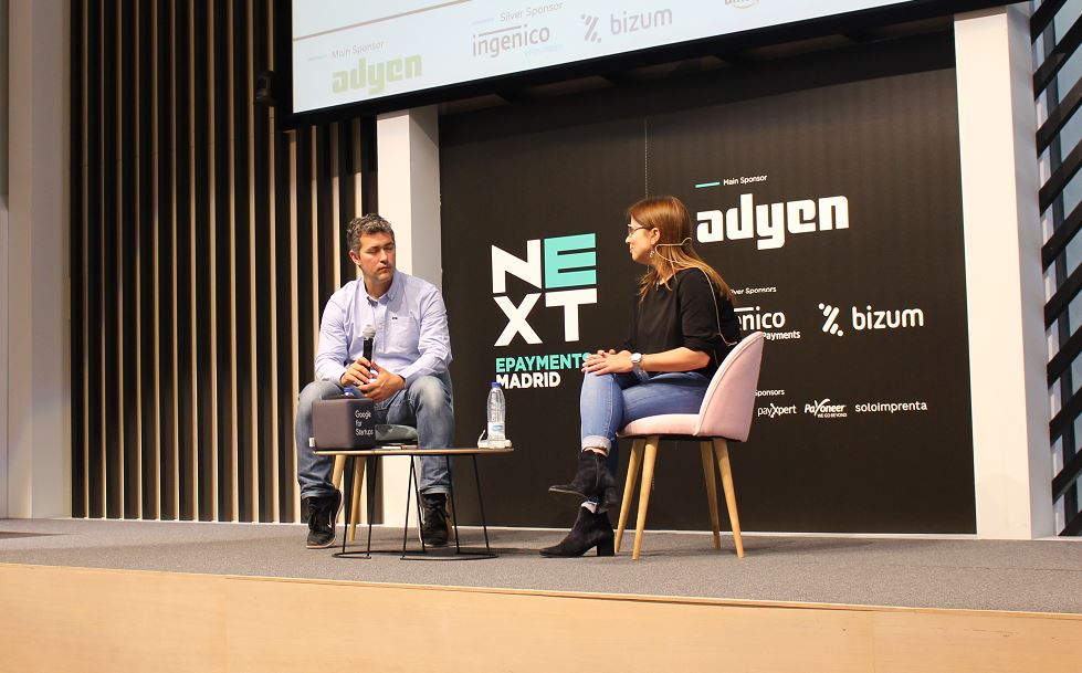 Dos personas sentadas con micr&oacute;fonos dialogan en un evento de pagos electr&oacute;nicos en Madrid. Fondo con logos de "NEXT" y patrocinadores: Adyen, Bizum, Ingenico.