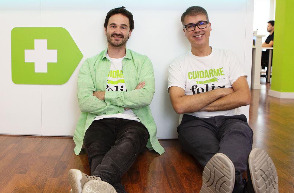 Dos personas sentadas en el suelo, sonriendo, con camisetas que dicen "CUIDARME" y "feliz", en una oficina moderna con suelos de madera. Logo de fondo.