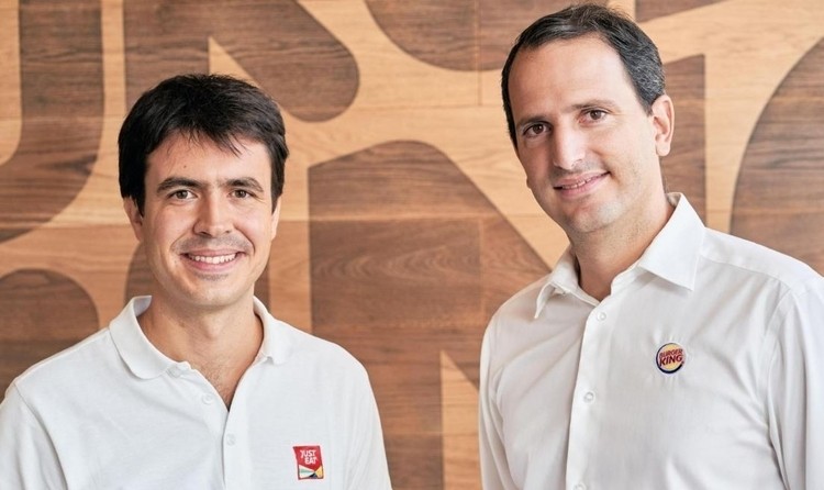 Dos hombres sonríen, vistiendo camisas blancas con logotipos de Just Eat y Burger King, en un fondo de madera corporativa.