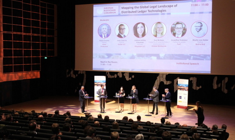 Personas de pie debaten en un escenario con proyector; público sentado observa. Texto: "Mapping the Global Legal Landscape of Distributed Ledger Technologies". Evento de conferencia.
