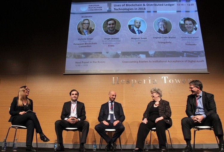Cinco personas sentadas en sillas, sonrientes, bajo una pantalla que muestra detalles de una convenci&oacute;n sobre blockchain en Hesperia Tower.