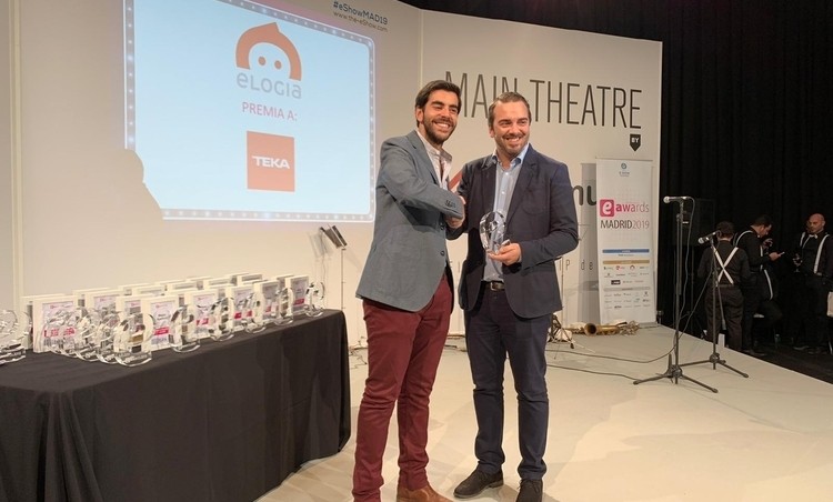 Dos personas posan en un escenario, una entrega un trofeo a la otra. A la izquierda, hay premios sobre una mesa y una pantalla proyecta "WIN THEATRE".