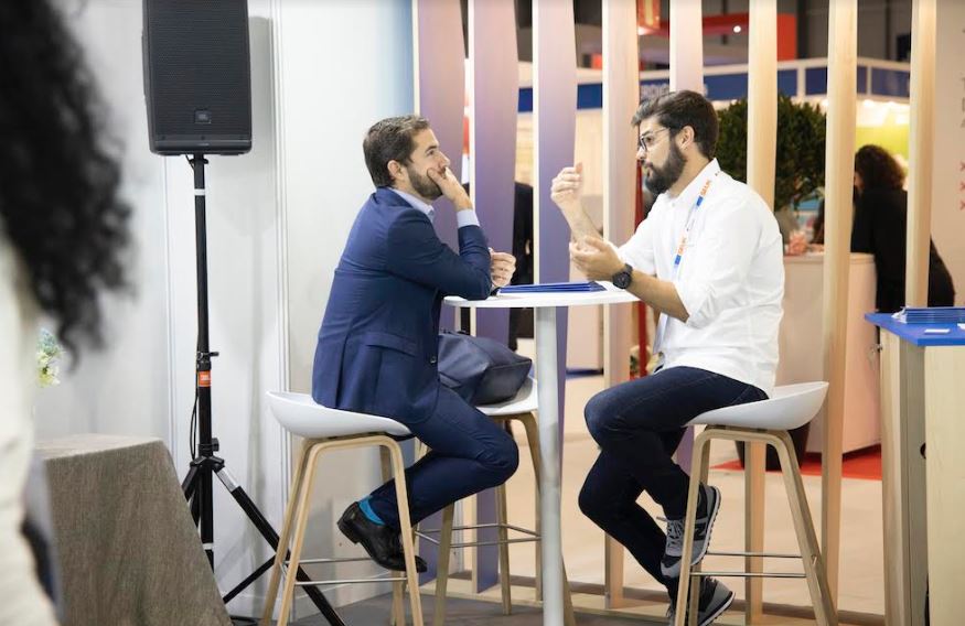 Dos hombres conversan animadamente mientras est&aacute;n sentados en taburetes blancos alrededor de una mesa alta en un entorno de exposici&oacute;n profesional.