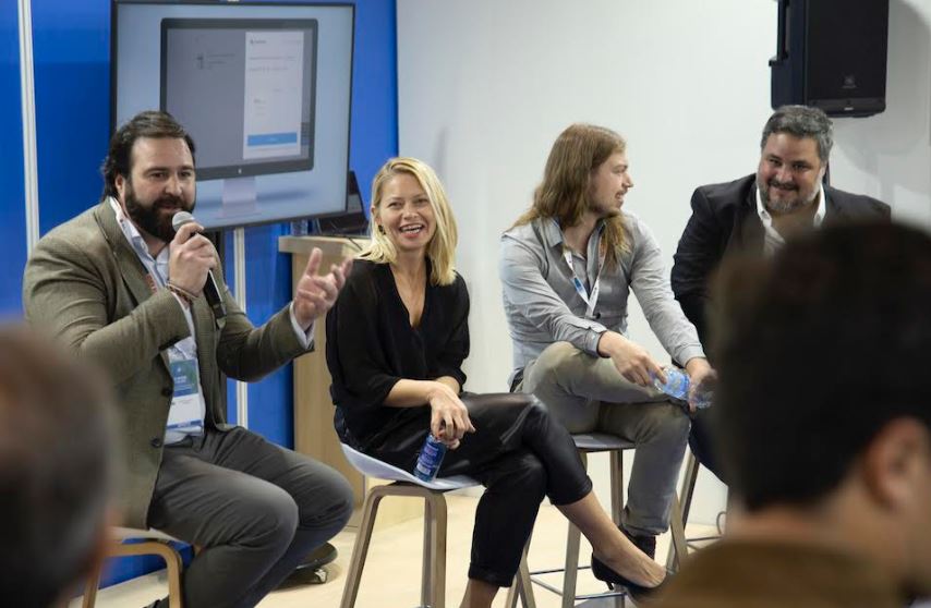 Personas sentadas conversando en un panel, cada una con micr&oacute;fono o botella de agua; pantalla de fondo muestra software. Ambiente profesional y din&aacute;mico.