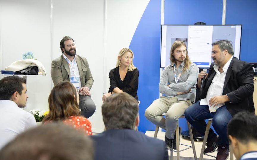 Panelistas dialogando en un evento empresarial, rodeados de asistentes. Pantalla detr&aacute;s con diapositiva visible. Ambiente de networking profesional.