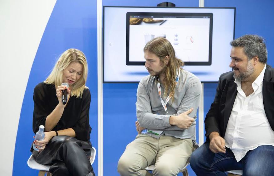 Mujer hablando en un micr&oacute;fono, sentada junto a dos hombres, con pantalla de fondo. Ambiente de conferencia. Potencial networking y di&aacute;logo profesional.