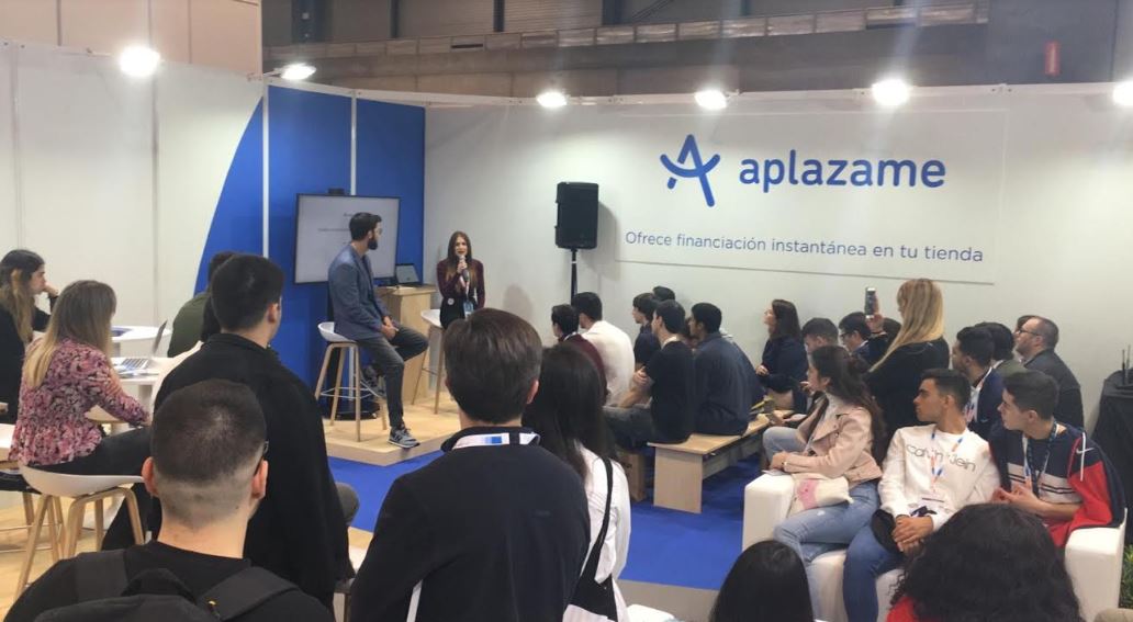 Persona hablando a audiencia sentada en un evento. Cartel: "aplazame, Ofrece financiaci&oacute;n instant&aacute;nea en tu tienda". Ambiente de feria o exposici&oacute;n.