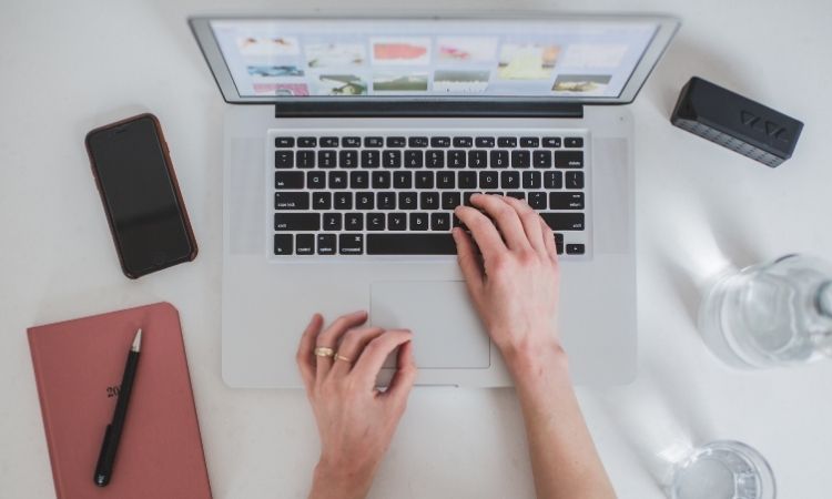 Manos tecleando en un portátil sobre un escritorio con móvil, cuaderno y vaso de agua; contexto de trabajo digital, productividad y tecnología.