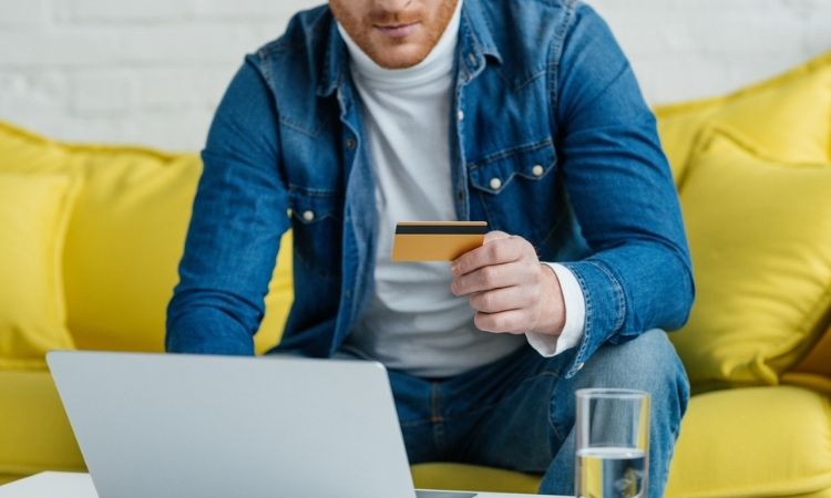 Persona sujeta una tarjeta de crédito frente a un portátil sobre una mesa; contexto de compra online en un salón con sofá amarillo.