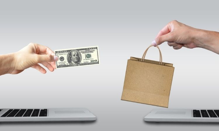 Dos manos intercambian billete y bolsa de papel sobre ordenadores portátiles en fondo blanco, simbolizando el comercio electrónico y la compra online.