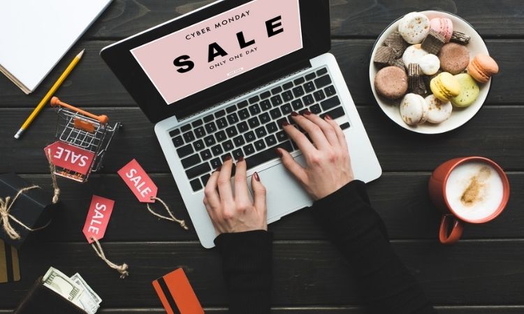 Manos tecleando en un portátil con anuncio de "CYBER MONDAY SALE ONLY ONE DAY". Alrededor, taza de café, macarons, carro de compras y dinero.