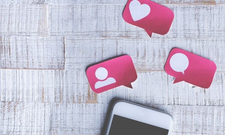 Smartphone sobre mesa de madera con íconos de redes sociales flotantes: usuario, corazón y mensaje. Conexión digital y engagement visual destacados.
