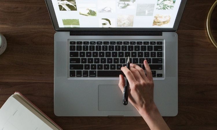 Mano escribe en portátil sobre mesa de madera; pantalla muestra imágenes variadas. Contexto de trabajo creativo o marketing digital. Cuaderno cercano.