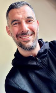 Hombre sonriente con barba y cabello corto, lleva puesta una chaqueta negra. Fondo claro y suave.