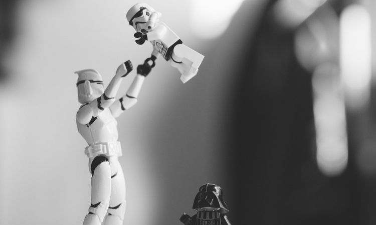 Muñeco Stormtrooper levanta a otro pequeño; figura de Darth Vader observa. Entorno desenfocado. Ideal para marketing juguetería o coleccionismo.