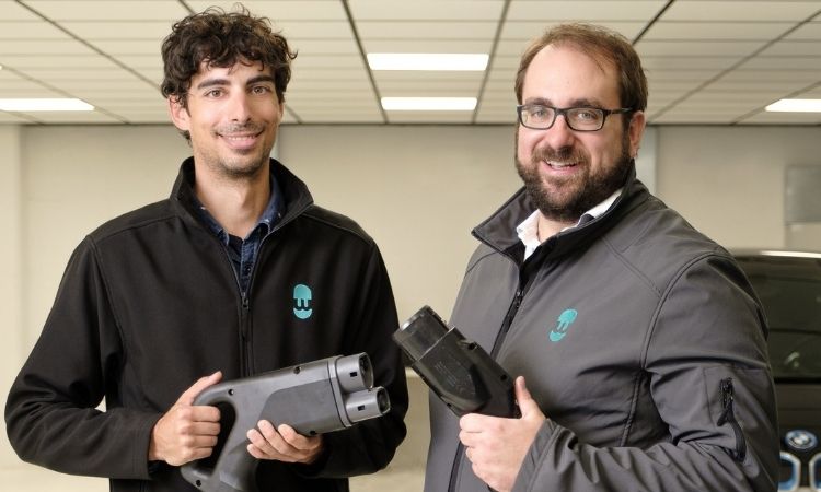Dos hombres sostienen conectores de carga de veh&iacute;culos el&eacute;ctricos en un entorno de sala, destacando la innovaci&oacute;n y sostenibilidad en movilidad el&eacute;ctrica.