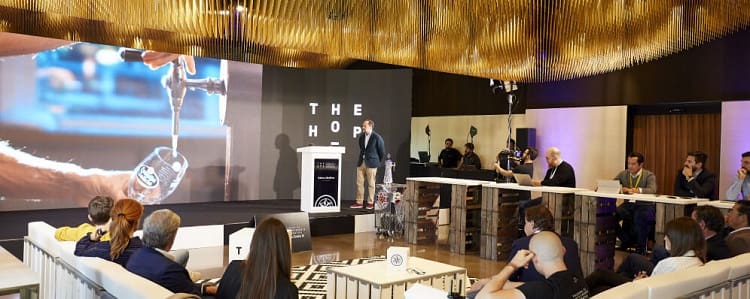 Persona presenta en escenario con pantalla grande mostrando vaso bajo grifo de cerveza; "The Hop" texto visible; audiencia atenta en sala moderna.