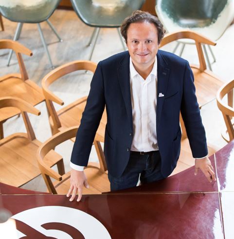 Un hombre sonriente, de pie y apoyado en una mesa con s&iacute;mbolo blanco; varias sillas de madera y metal en un entorno de oficina moderno.