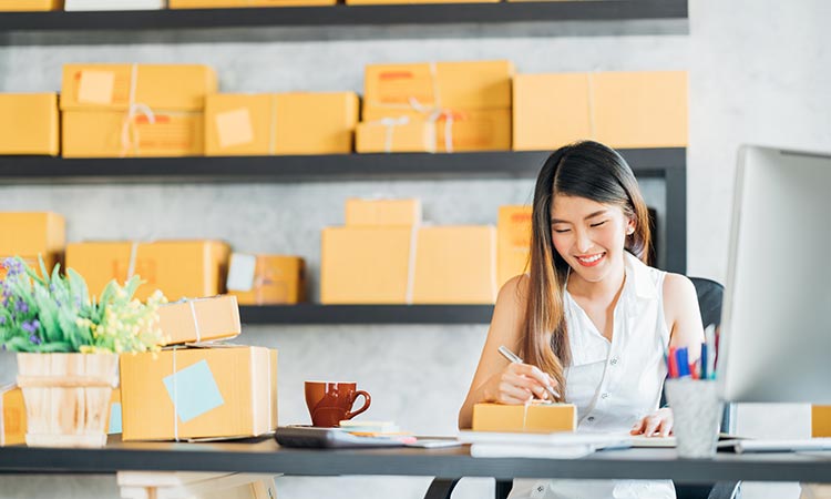 Mujer sonriente, escribiendo sobre paquetes en una oficina con cajas apiladas en estanterías y flores sobre el escritorio. Marketing de envíos.