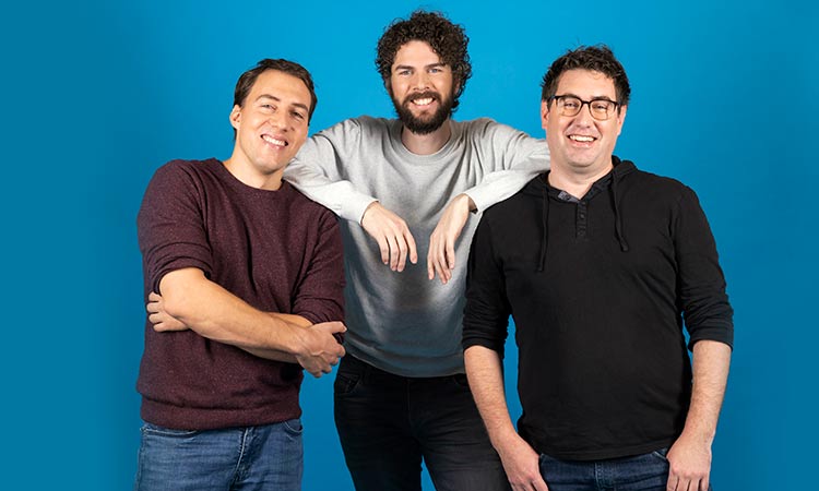 Tres personas, de pie y sonrientes, posan contra un fondo azul, transmitiendo trabajo en equipo y camarader&iacute;a.