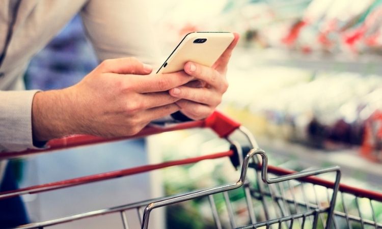 Persona usa móvil mientras empuja carrito en supermercado, rodeado de estantes llenos de productos. Contexto moderno y digitalización de compras.