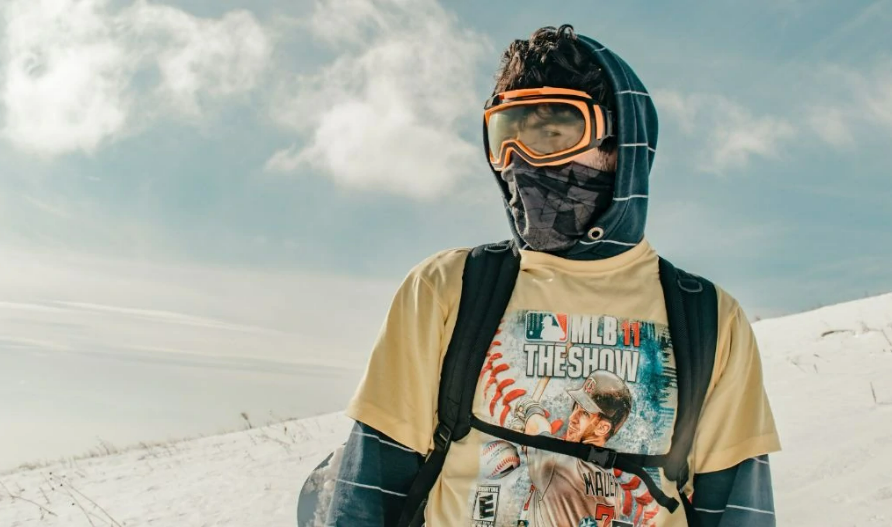 Persona con gafas de nieve camina en una colina nevada. Viste camiseta con texto: "MLB 11 THE SHOW" y una imagen de b&eacute;isbol. Cielo azul.