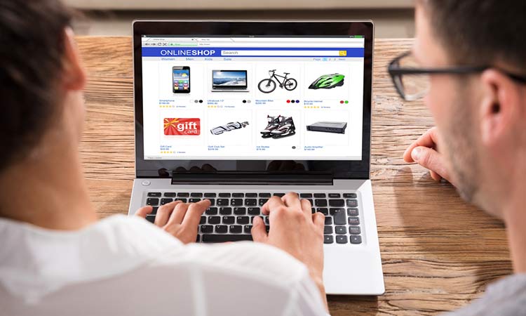 Pantalla de portátil muestra una tienda online, dos personas mirando productos electrónicos y regalos desde una mesa de madera, destacando el e-commerce.