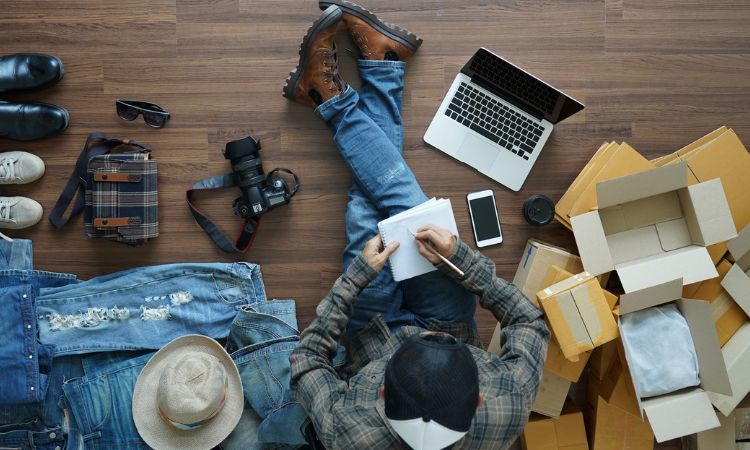 Persona escribiendo en un bloc, rodeada de ropa, zapatos y cajas, junto a un portátil y una cámara sobre suelo de madera. Palabras clave: e-commerce, preparación de pedidos.