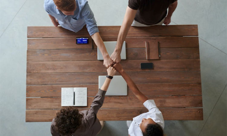 Manos unidas sobre mesa de madera, simbolizando colaboración. Alrededor, cuadernos, portátil y teléfono móvil. Ambiente de trabajo en equipo efectivo.
