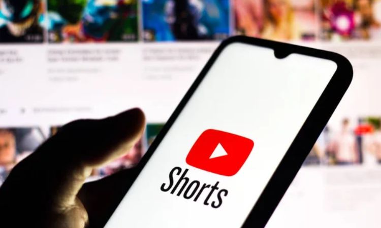 Teléfono mostrando el logo de YouTube Shorts; al fondo, una pantalla desenfocada de videos. Texto: "Shorts". Ambiente digital y multimedia.