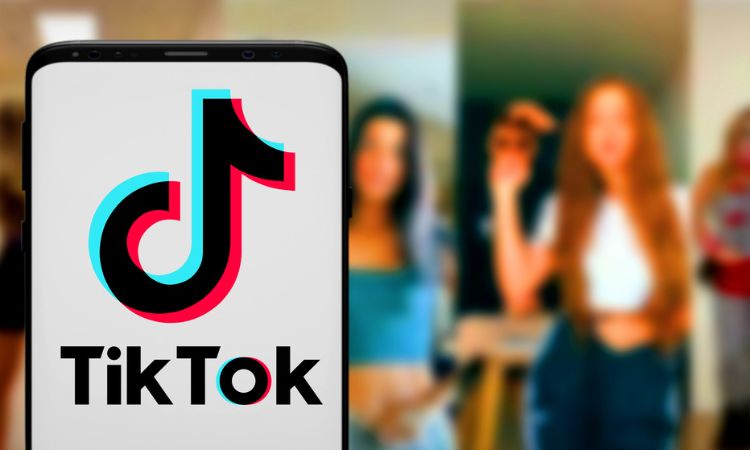 Teléfono muestra el logo de TikTok; al fondo, tres personas desenfocadas bailan en una habitación, transmitiendo creatividad y conexión social.