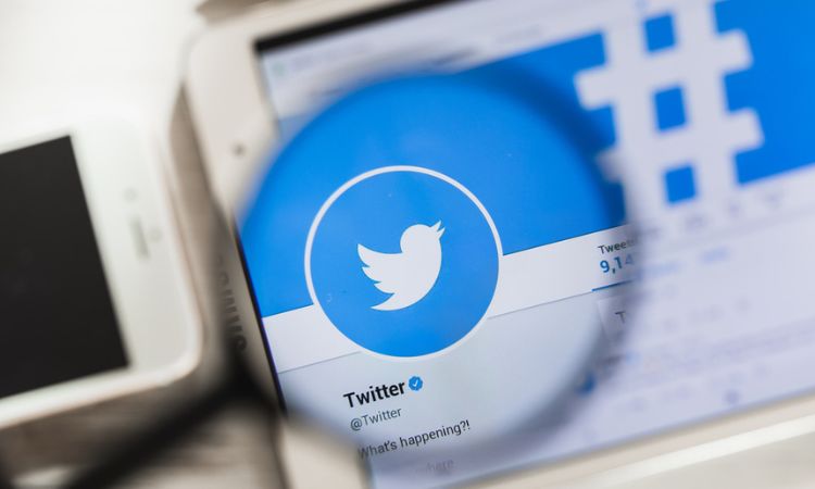 Logotipo de Twitter ampliado con lupa, pantalla de móvil en segundo plano, destacando interacción en redes sociales y comunicación digital.