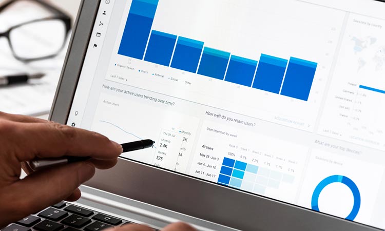 Pantalla de ordenador muestra gráficos de análisis; usuario señala con bolígrafo. Fondo de oficina. Texto visible: "How are your active users trending over time?". Marketing digital.