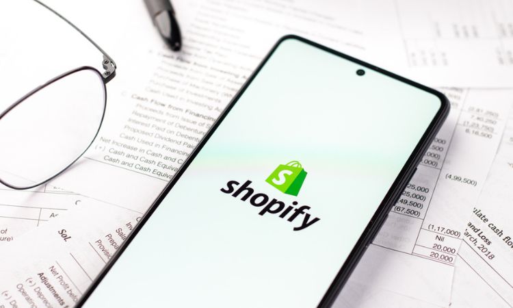 Un móvil con la aplicación de Shopify en la pantalla descansa sobre unos papeles financieros, junto unos lentes y un bolígrafo.