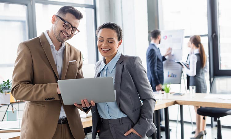Profesionales sonríen mientras miran un portátil en una oficina moderna; al fondo, colegas colaboran frente a un gráfico. Trabajo en equipo y tecnología.