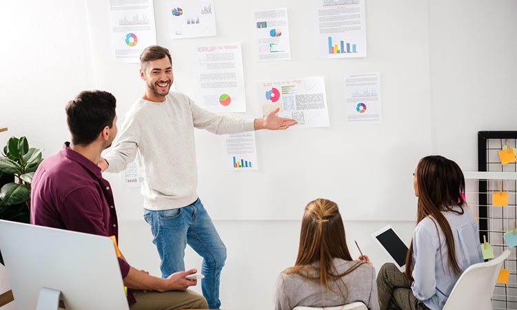 Hombre presenta gráficos en sala de reuniones; otros tres asistentes escuchan atentos, rodeados de informes en papel. Ambiente de colaboración empresarial.