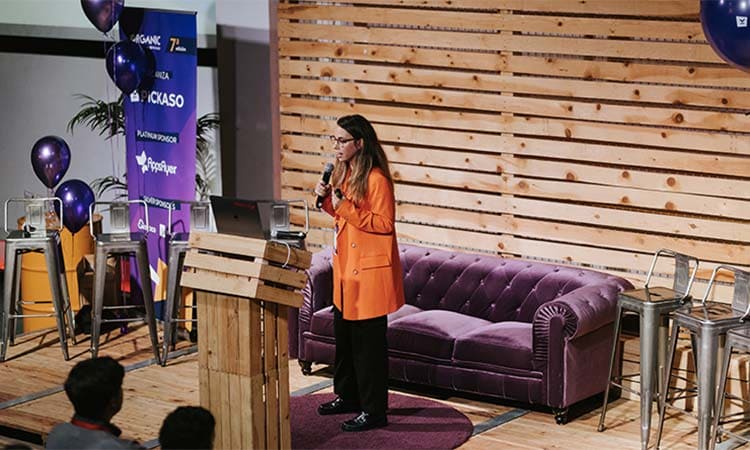 Persona hablando en escenario con micrófono, fondo de sofá morado y pared de madera, carteles con logos y globos; posible evento de networking.