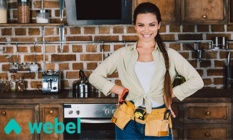 Mujer sonriente con cinturón de herramientas, de pie en una cocina rústica con pared de ladrillo. Logotipo "webel" en la esquina. Ideal para marketing de renovaciones.