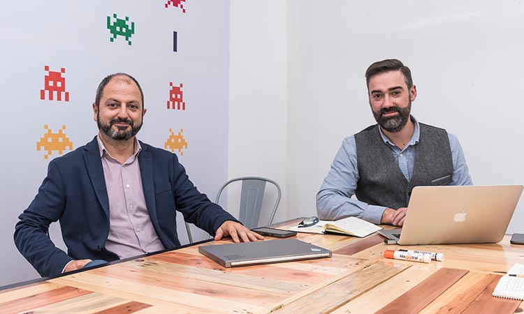 Dos hombres sentados, trabajando en escritorio con portátil y cuadernos. Fondo con gráficos de videojuegos. Entorno de oficina creativo y digital.