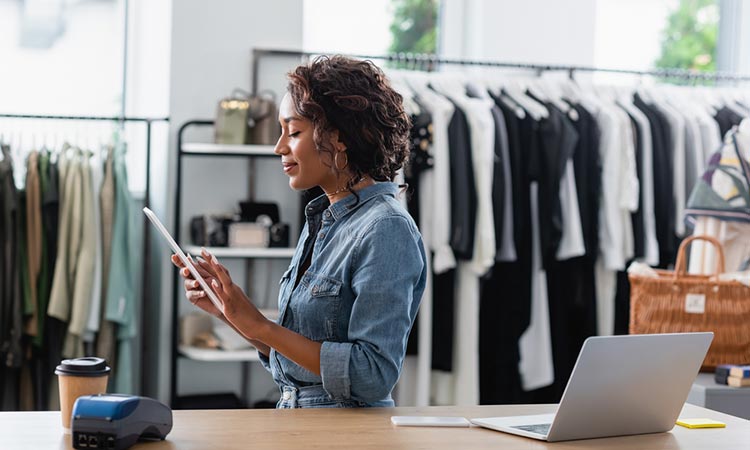 Mujer usando tableta en tienda de ropa, rodeada de percheros, portátil y lector de tarjetas. Ambiente profesional de moda y tecnología.