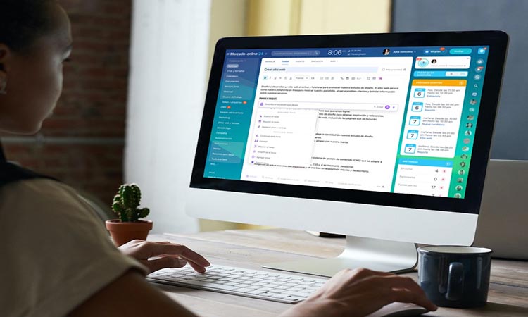 Persona trabajando en un ordenador de sobremesa revisando correos electrónicos; entorno de oficina moderno con una planta pequeña al lado del teclado.