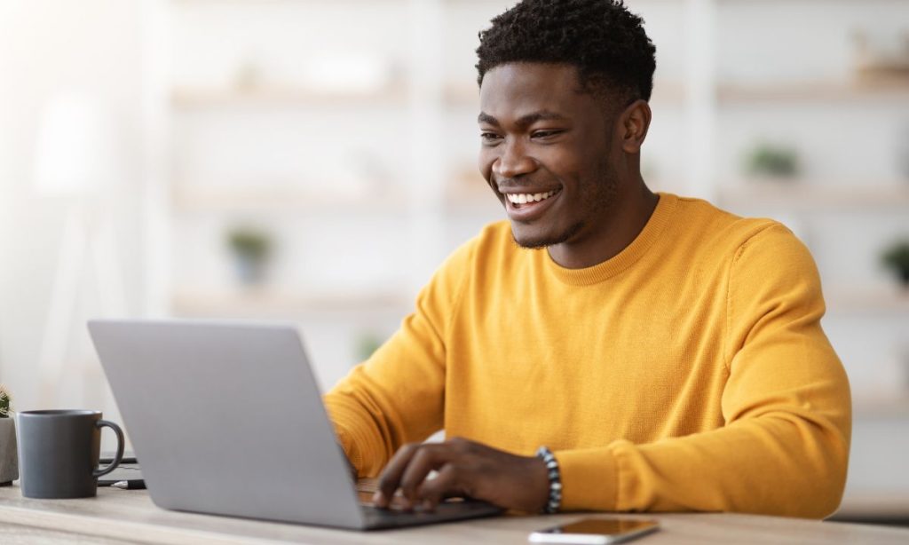 Imagen de un chico joven con un jersey amarillo frente al PC, sonriente, trabajando.