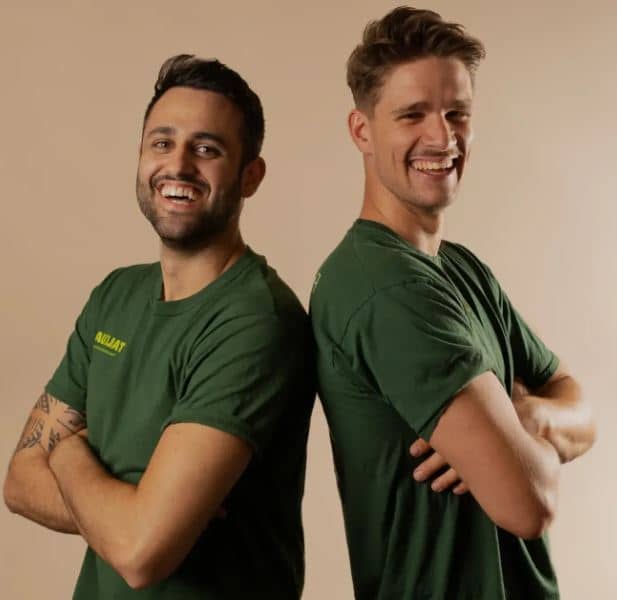 Dos hombres se apoyan de espaldas, cruzando los brazos y sonriendo, ambos con camisetas verdes y la palabra "ABUANT" en una de ellas. Entorno beige.