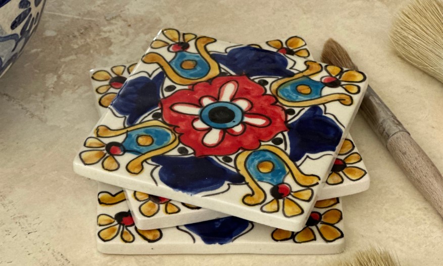 Azulejos decorativos apilados exhiben dise&ntilde;os vibrantes florales en azul y rojo; al lado, un pincel sobre una superficie clara. Decoraci&oacute;n artesanal.