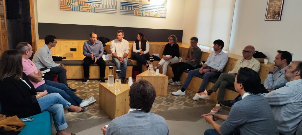 Personas sentadas en c&iacute;rculo, dialogando en una sala moderna, con mesas de madera al centro. Ambiente colaborativo para networking y aprendizaje.
