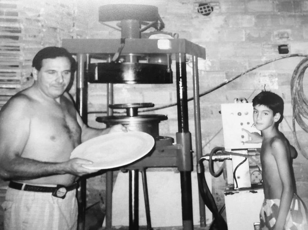 Hombre sostiene una placa, ni&ntilde;o junto a m&aacute;quina industrial en taller de ladrillos. Ambiente vintage refleja tradici&oacute;n y manualidad.