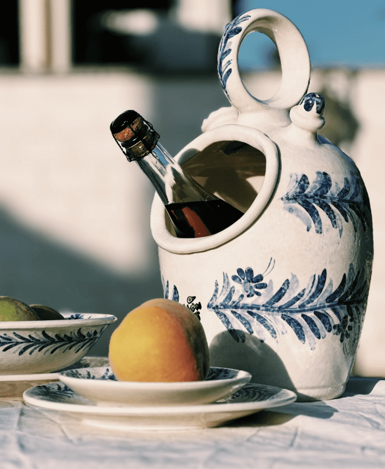 Botella inclinada en jarra cer&aacute;mica decorada junto a plato con melocot&oacute;n, en mesa exterior soleada. Perfecto para promoci&oacute;n de decoraci&oacute;n y estilo.
