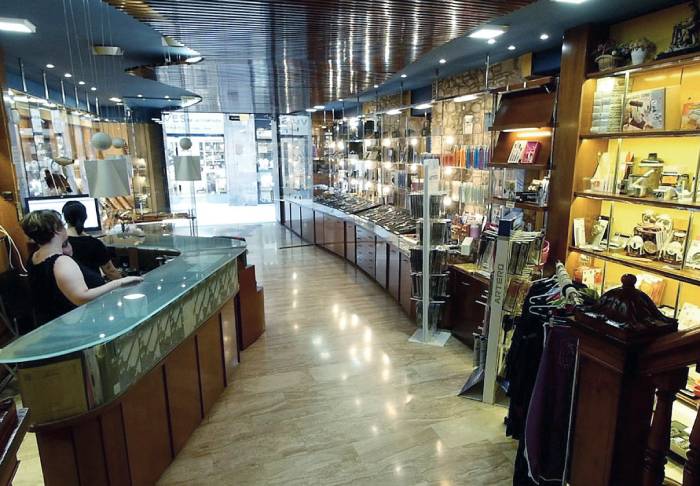 Tienda de regalos; dos personas esperan en el mostrador de cristal curvo; vitrinas iluminadas muestran art&iacute;culos diversos; ambiente elegante y moderno.
