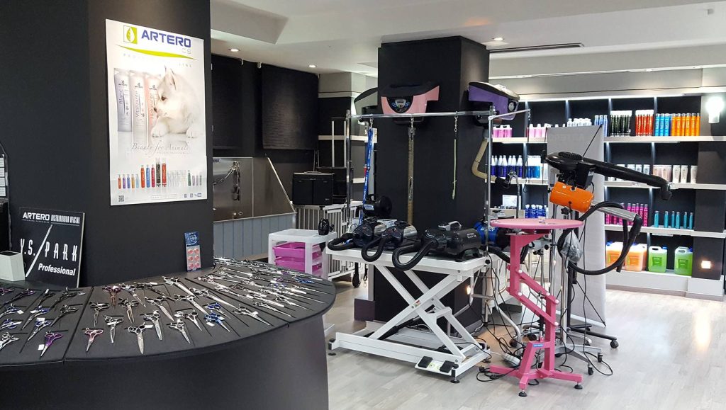 Tienda de est&eacute;tica canina con tijeras expuestas sobre mostrador curvo; fondo con productos, mesas de trabajo y carteles publicitarios de "ARTERO".